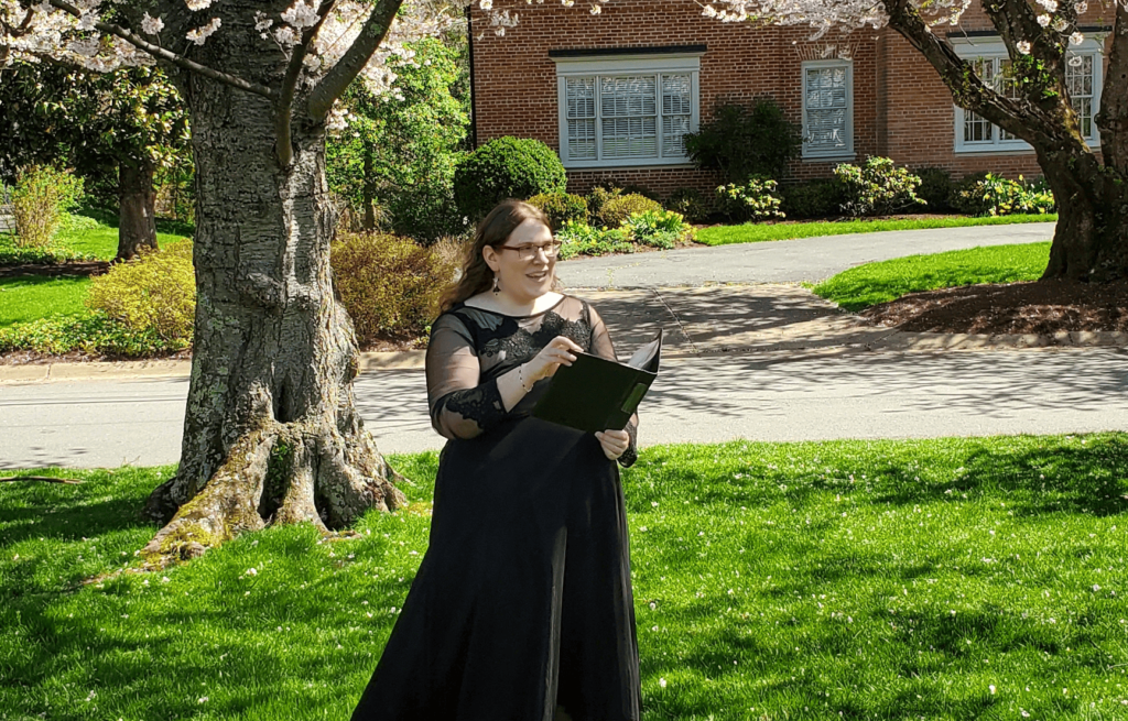 Emily Karp standing with a binder in her hands, officiating a funeral under cherry blossom trees.