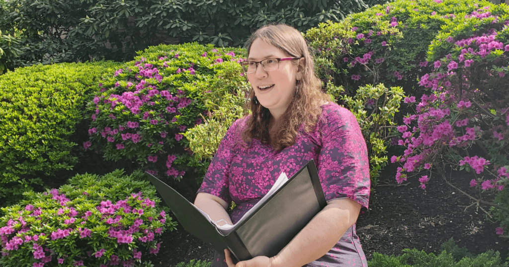 Image of Emily Karp, a white woman with light brown curly hair wearing a dress that's navy blue with many hot pink flowers on the top part of the dress and they fade as they go down the dress to less and less hot pink flowers over top of the blue. She stands in front of Green bushes with pink flowers on them, while she holds a binder and reads aloud from it to an unseen audience.